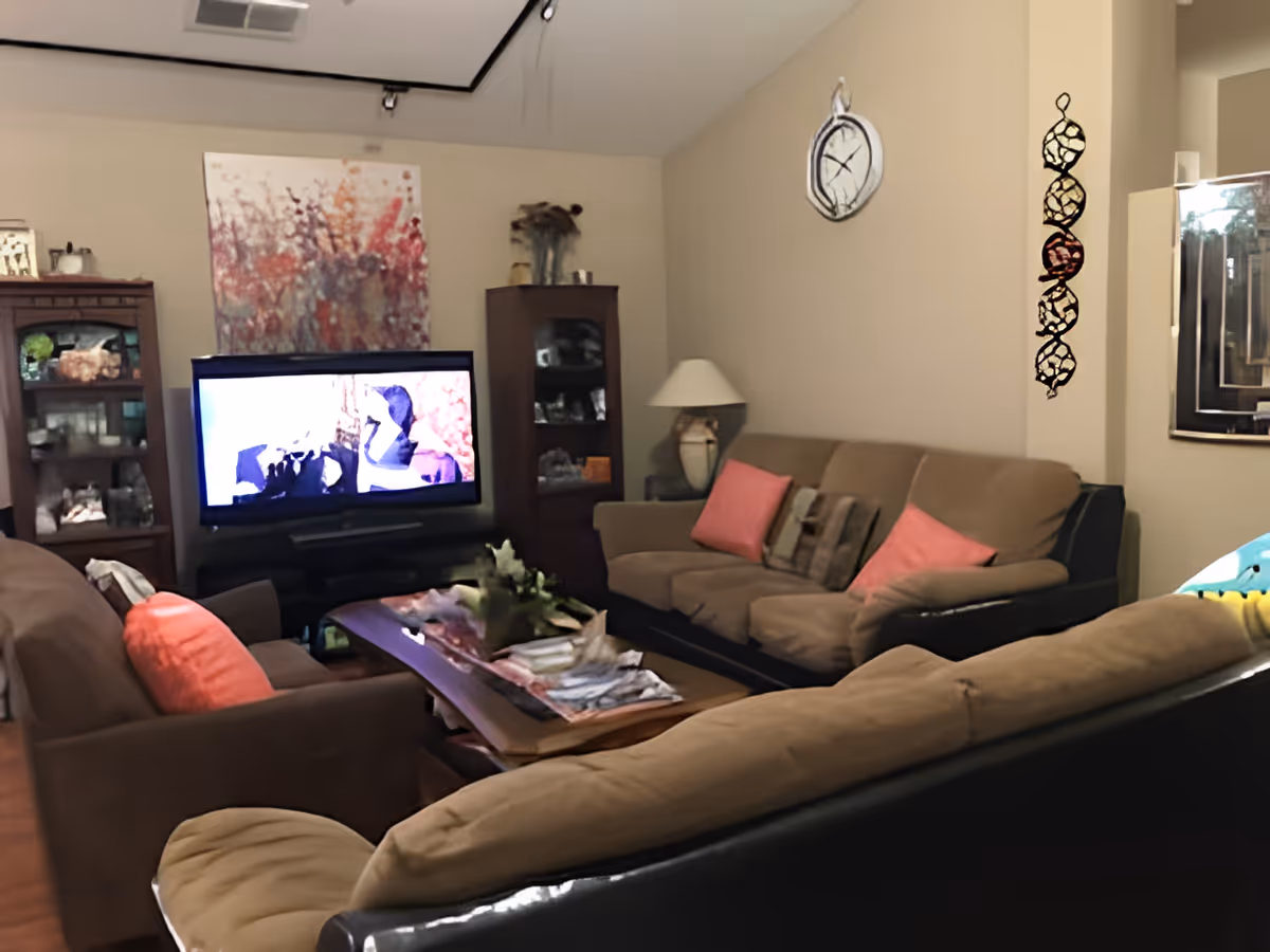 A cozy living room with three brown sofas adorned with pink and patterned cushions arranged around a wooden coffee table with magazines and a decorative plant. A flat-screen TV is on a stand against the wall, flanked by two wooden display cabinets with glass doors containing various decorative items. The walls are beige with a large abstract painting behind the TV, a wall clock, a lamp on a side table, and a decorative hanging piece. A mirror is partially visible on the right side.