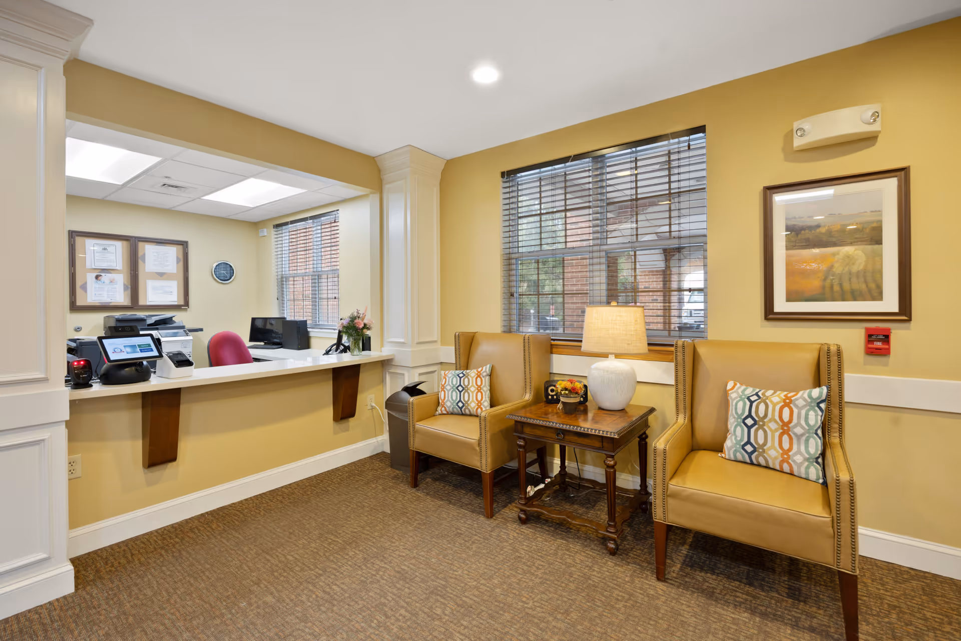 Reception area with a counter and office equipment behind it, two tan armchairs with patterned cushions, a wooden side table with a lamp and decorative items, a window with blinds, and framed artwork on the wall.