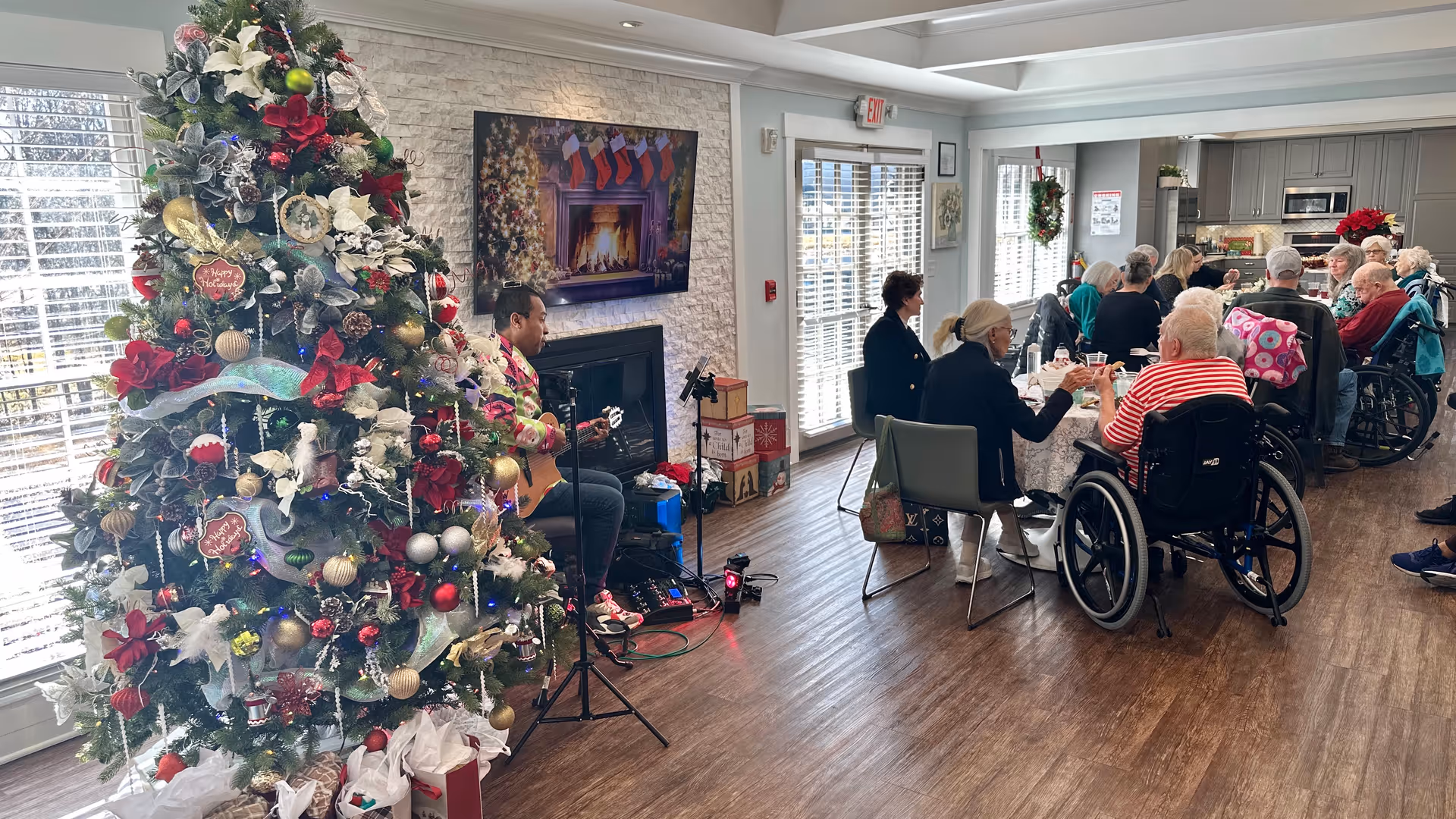 A festive common room with a large decorated Christmas tree and seniors seated at tables, some in wheelchairs, while a musician plays guitar.