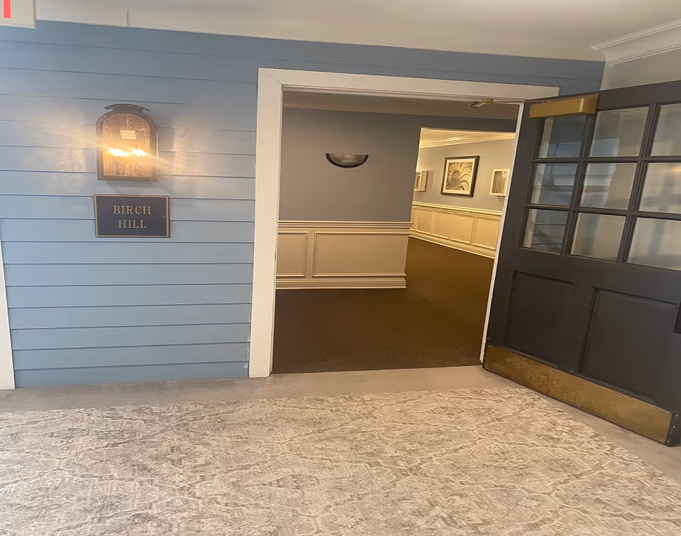 An interior hallway entrance with a blue wall on the left featuring a lit wall sconce above a sign that reads 'BIRCH HILL'. To the right, there is a dark-colored door with glass panes propped open, leading into a carpeted hallway with framed artwork on the walls.