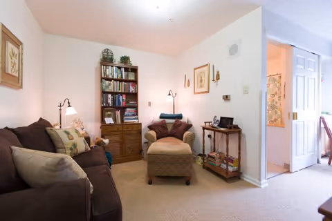 A cozy living room with a brown sofa adorned with pillows on the left, a cushioned armchair with an ottoman in the center, and a wooden bookshelf filled with books against the back wall. There is a small wooden side table with books and a digital photo frame on the right. The room is softly lit with wall sconces and a floor lamp. An open doorway on the right leads to a bathroom with a floral shower curtain.