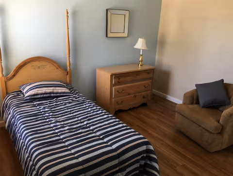 A bedroom with a wooden single bed featuring a striped blue and white bedspread and matching pillow. Next to the bed is a wooden dresser with three drawers and a small lamp on top. A cushioned armchair with a dark pillow is positioned against the wall. The floor is wooden, and a small square mirror hangs above the dresser.