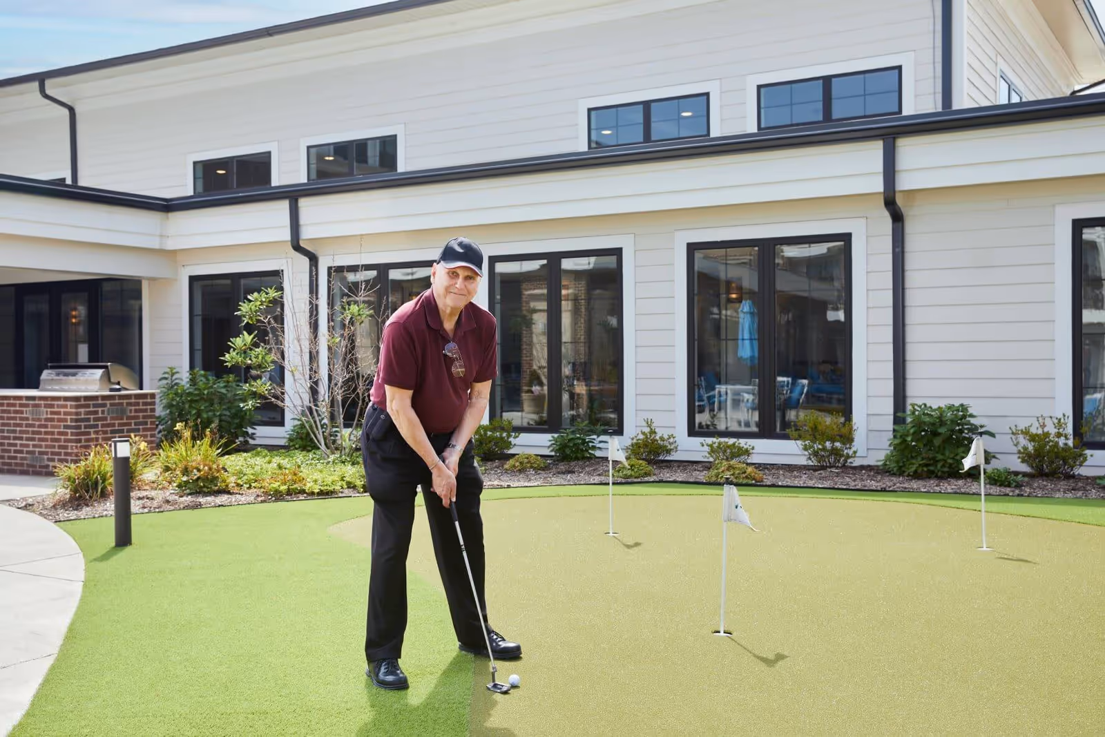 An elderly man wearing a maroon polo shirt, black pants, and a black cap is playing putting golf on a small artificial green area outside a senior living facility building with large windows and some landscaping.