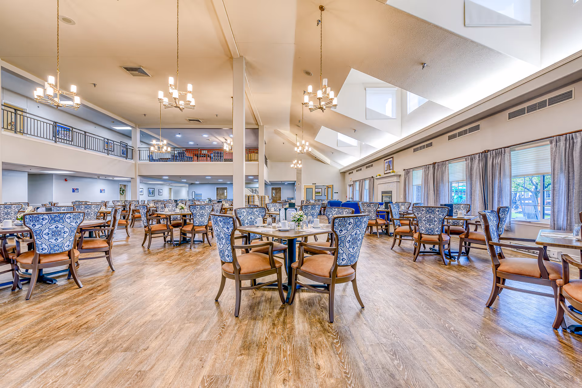 Spacious senior living dining room with multiple wooden tables and chairs featuring blue and white patterned backs and brown seats. The room has high ceilings with modern chandeliers, large windows with curtains letting in natural light, and a fireplace along one wall. There is a second-floor balcony overlooking the dining area.