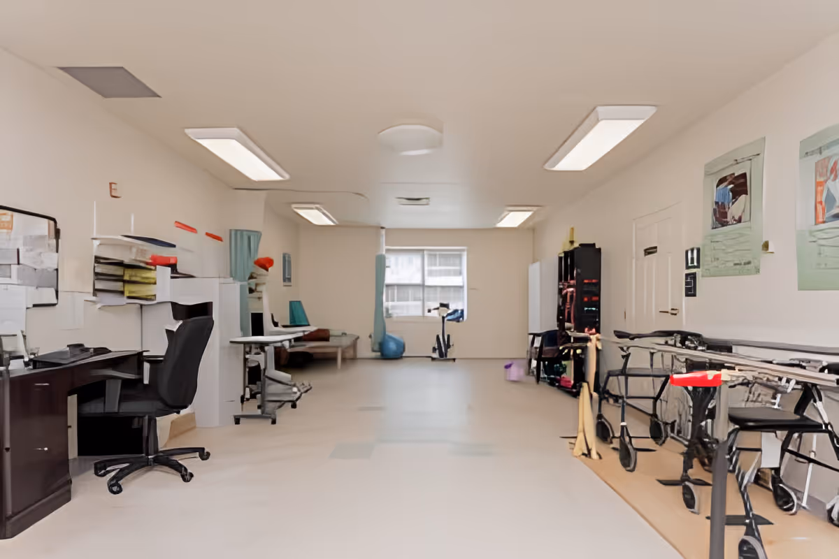 A large multipurpose interior room with desks and office chairs on the left, mobility walkers and equipment lined up on the right, and exercise/therapy gear toward the back.