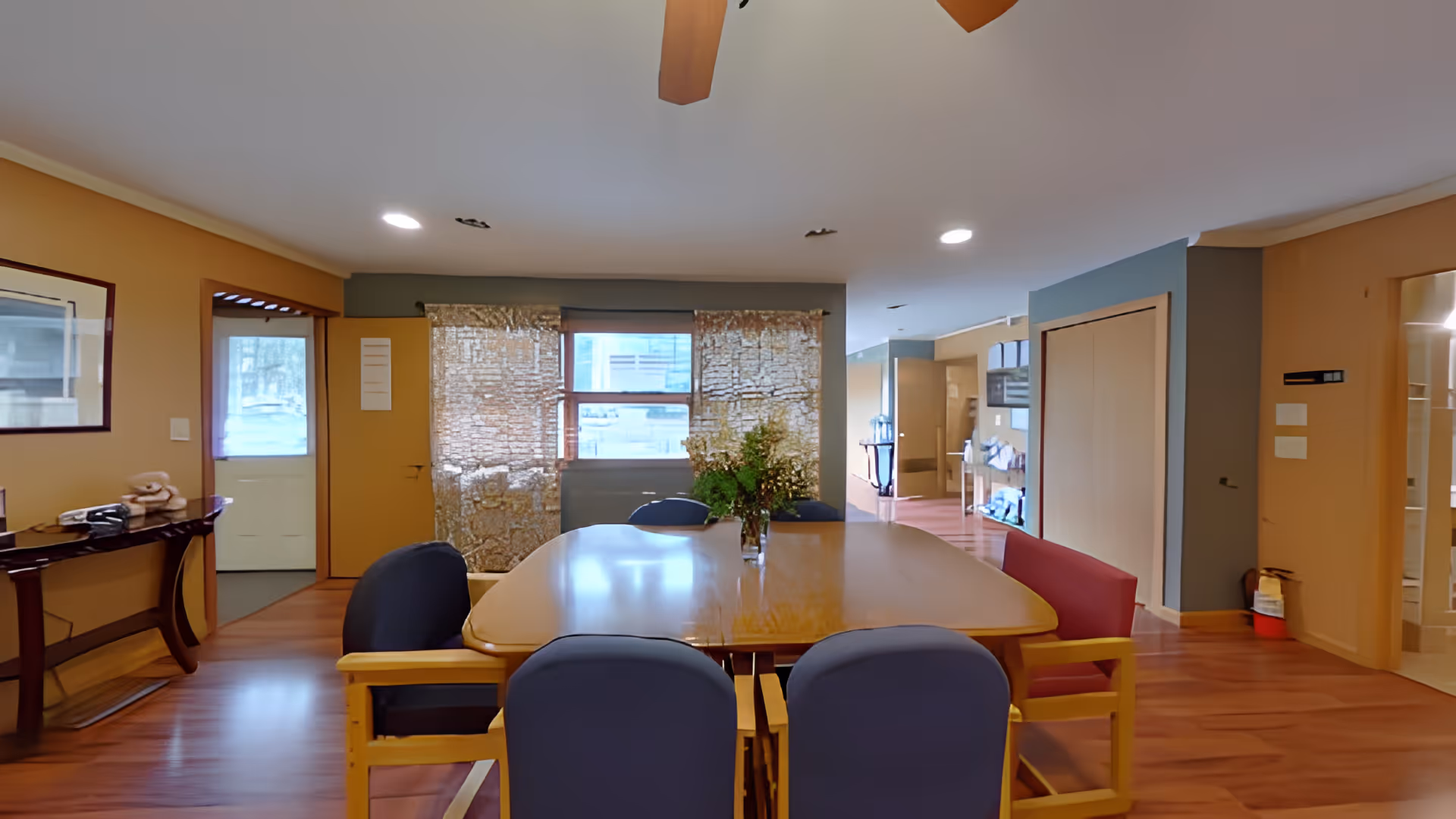 Communal dining area with a large wooden table surrounded by chairs, windows, and warm-toned walls.