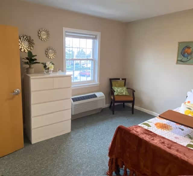 A small bedroom with a bed covered in a floral quilt and a rust-colored throw blanket. There is a wooden chair with a green cushion in the corner next to a window with a white frame. Below the window is a wall-mounted air conditioning unit. A white chest of drawers with decorative items on top is positioned against the wall near the door. The walls are beige, and there are three decorative round mirrors on the wall above the chest of drawers.