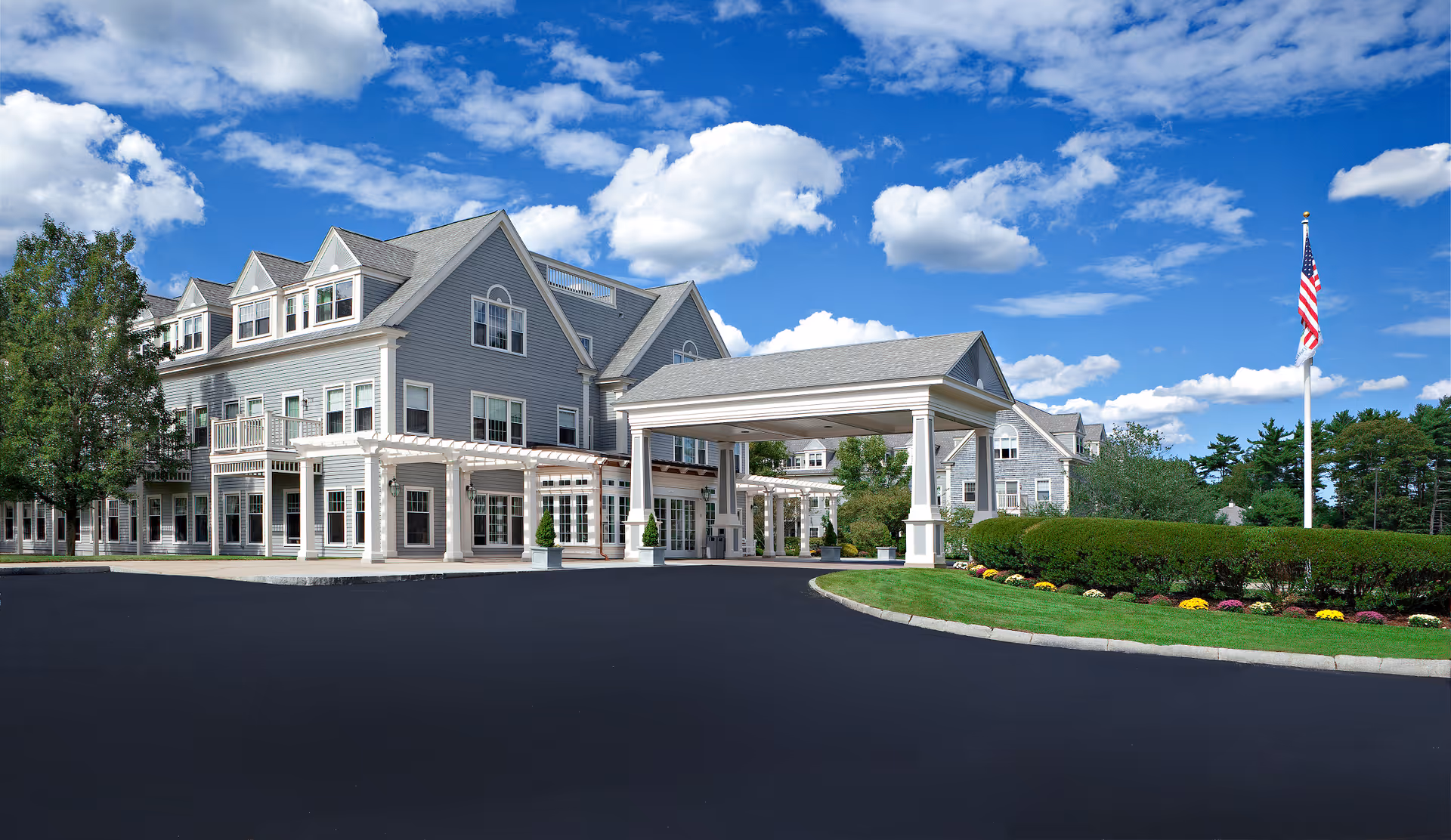 Exterior view of a large, multi-story senior living facility building with gray siding and white trim under a bright blue sky with scattered clouds. The building features multiple windows, a covered entrance with columns, and a well-maintained driveway and landscaped lawn with flowers and an American flag on a flagpole.