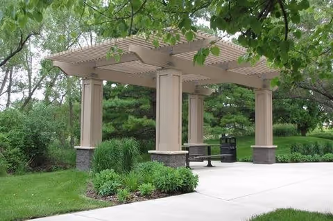 A beige pergola structure with four columns and a slatted roof situated on a concrete pathway surrounded by green grass, bushes, and trees in a landscaped outdoor area.