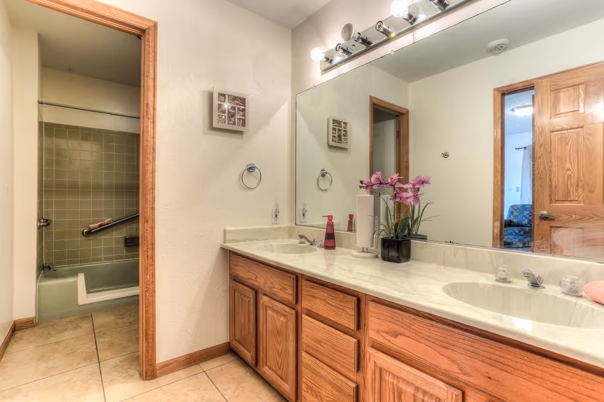 Bathroom with a double sink vanity featuring wooden cabinets and a large mirror above. There is a pink soap dispenser and a potted plant with purple flowers on the countertop. The bathroom has beige tiled floors and a doorway leading to a bathtub with green tiles and a grab bar.