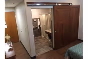 Interior view of a senior living facility room at Schowalter Villa showing a partial bedroom with a bed on the right, a wooden sliding door leading to a bathroom with a shower curtain, sink, and mirror, and a hallway with a wooden door and a small table with a flower vase on the left.