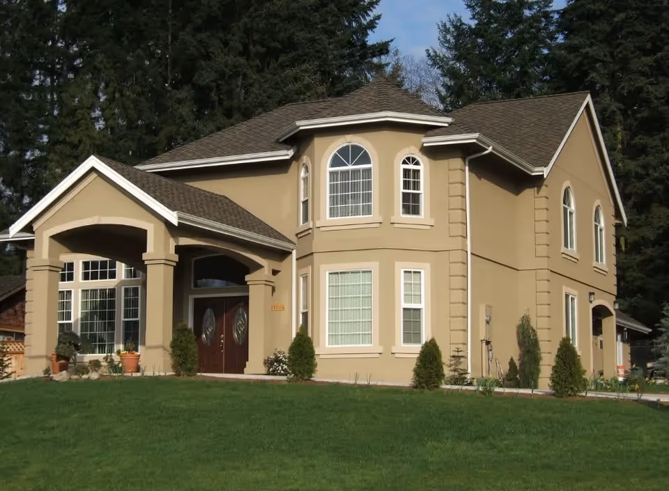 Exterior view of a two-story beige house with multiple windows, a covered front porch, and a well-maintained green lawn in front. Tall evergreen trees are visible in the background.