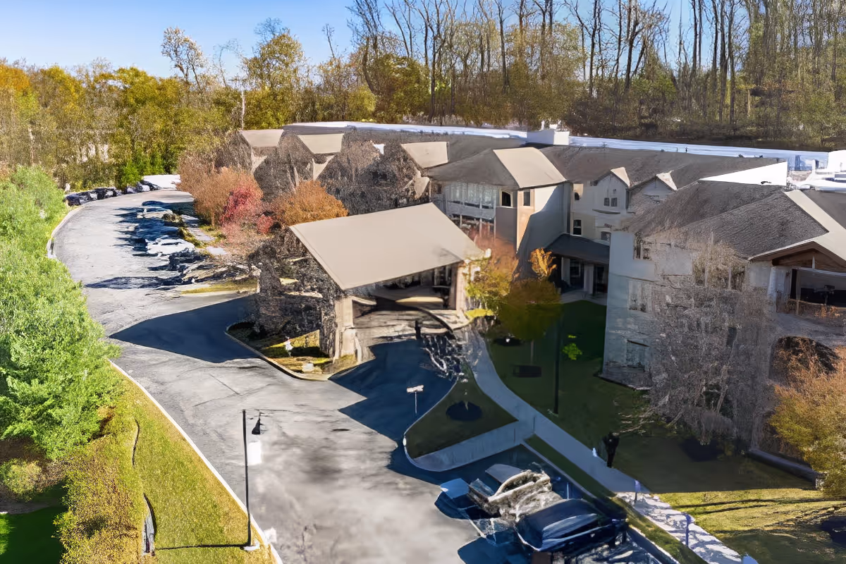 Aerial view of a senior living facility named Spring Mill, showing a large building complex surrounded by trees and greenery. There is a driveway leading to a covered entrance with parked cars nearby. The background includes a wooded area with tall trees.