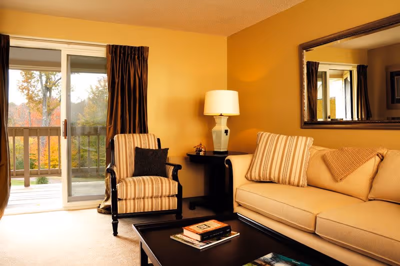A cozy living room with a beige sofa adorned with striped and solid pillows, a striped armchair with a dark cushion, a black coffee table with books on it, a side table with a lamp, and a large mirror on the wall. There is a sliding glass door with brown curtains leading to an outdoor balcony with trees visible outside.