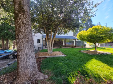 A grassy yard with trees and a covered patio attached to a light-colored two-story building.