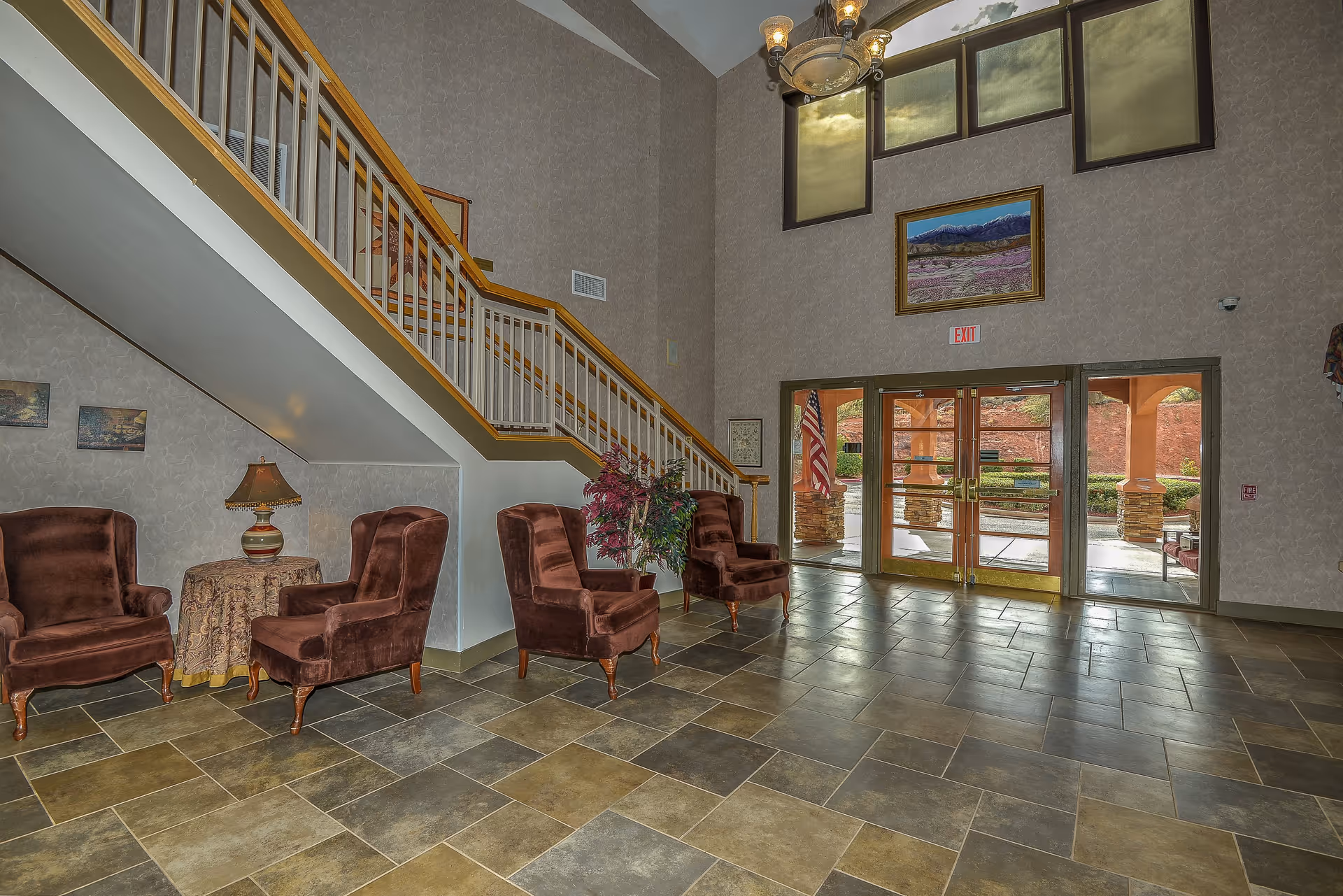 Interior view of a spacious assisted living community lobby with a tiled floor, four brown upholstered armchairs arranged in pairs near a staircase, a small round table with a decorative lamp, a potted plant, large windows above the entrance doors, and a framed landscape painting on the wall.