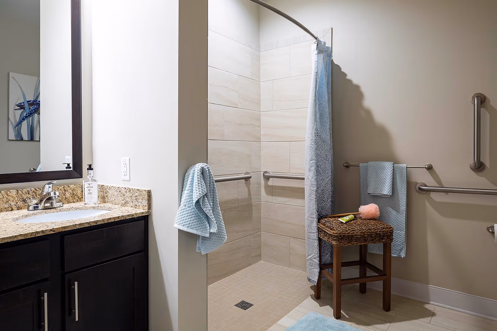 A bathroom with a granite countertop sink on the left, a large mirror above it, and a walk-in tiled shower with grab bars and a blue patterned shower curtain. There are blue towels hanging on the wall and a wicker stool with bath items on it inside the shower area.