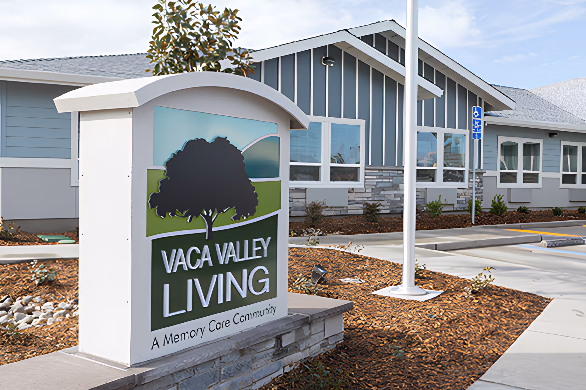 Sign reading 'Vaca Valley Living — A Memory Care Community' stands in landscaped grounds in front of the facility's blue-gray building.