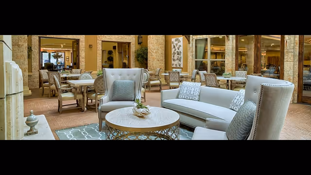 A cozy outdoor patio area with cushioned armchairs and a sofa arranged around a round coffee table on a patterned rug. In the background, there are dining tables and chairs set on a brick floor, with large windows and doors leading to an indoor space.
