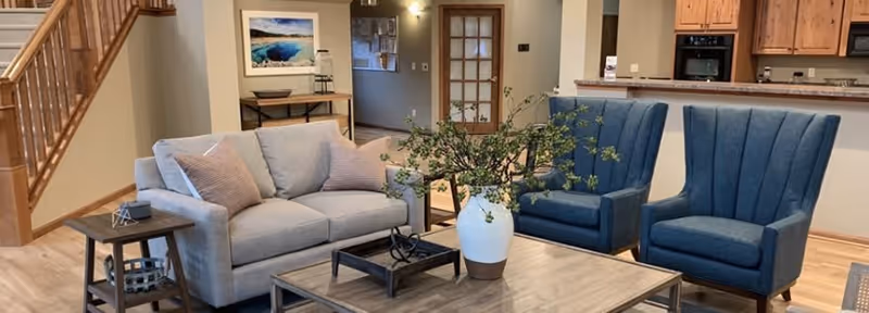 Cozy communal living area with a sofa, two blue armchairs, a coffee table with a vase of greenery, and an open kitchen in the background.