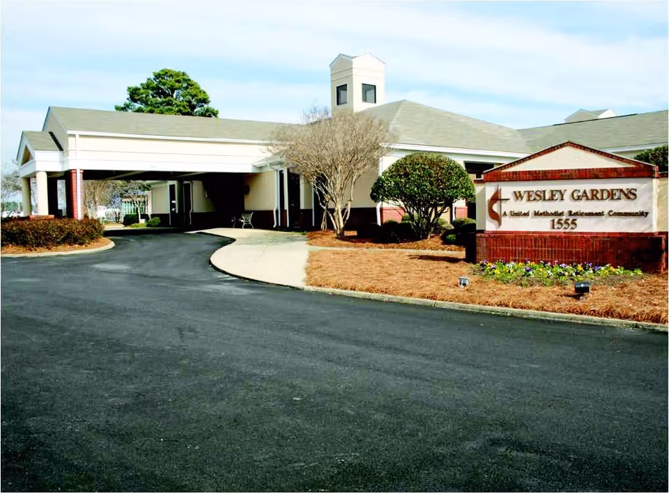 Exterior view of Wesley Gardens, a United Methodist Retirement Community building with a covered driveway entrance, landscaped bushes, and a sign displaying the community name and address 1555.