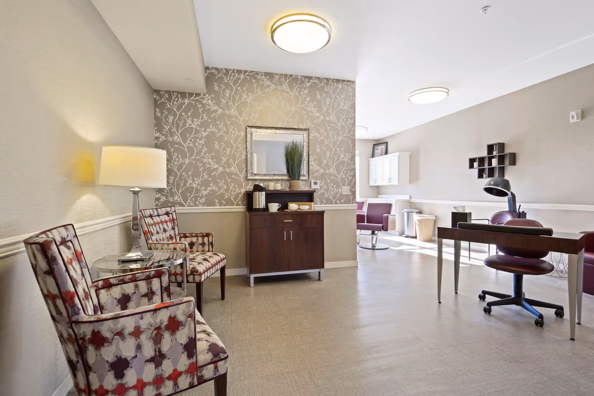A bright, modern interior room with patterned armchairs and a glass side table on the left, a wooden cabinet with a coffee pot and cups against a wallpapered wall in the center, and a desk with a rolling chair and lamp on the right. The room has light-colored walls and flooring, with additional seating and cabinetry visible in the background.