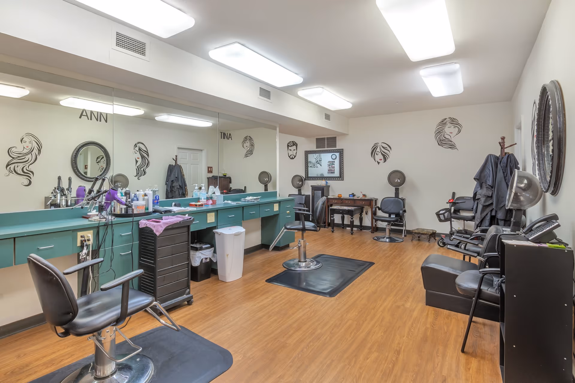 Well-lit hair salon with styling chairs, mirrors, countertops, and hair-drying stations in a senior living facility.