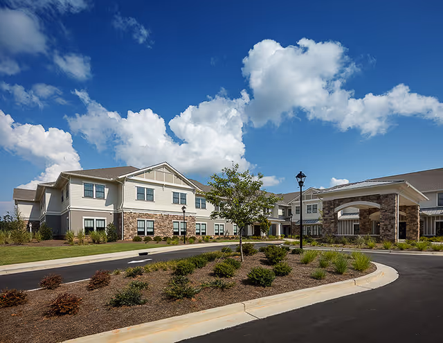 Exterior view of a senior living facility building with a stone and siding facade under a blue sky with scattered clouds. The driveway curves around landscaped areas with small bushes and a young tree, leading to a covered entrance supported by stone pillars.