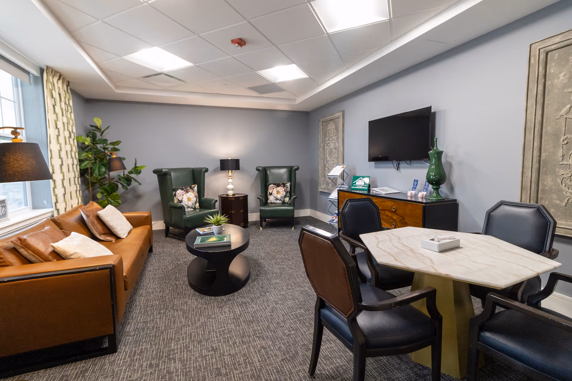 Comfortable lounge with a leather sofa, two green wingback chairs, a round coffee table, a small dining table with chairs, and a wall-mounted TV.