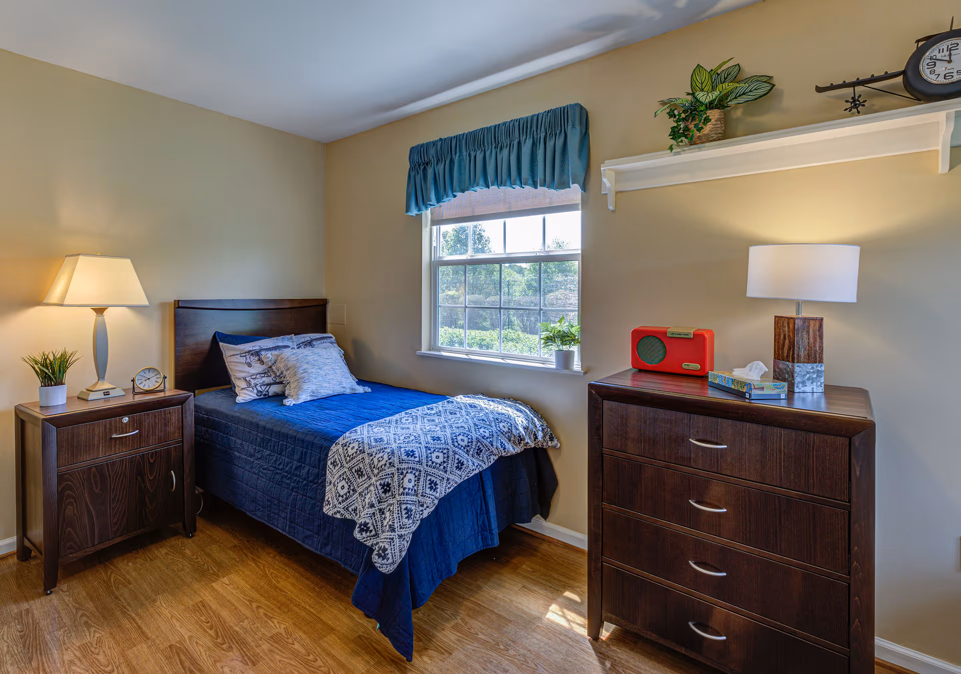 A cozy bedroom with a single bed covered in a blue quilt and patterned blanket. There are two pillows on the bed. Next to the bed is a wooden nightstand with a lamp, a small plant, and a clock. A window with a blue valance lets in natural light. On the opposite side of the bed is a wooden dresser with a lamp, a red radio, a tissue box, and a small plant on a white shelf above it. The room has light-colored walls and wooden flooring.