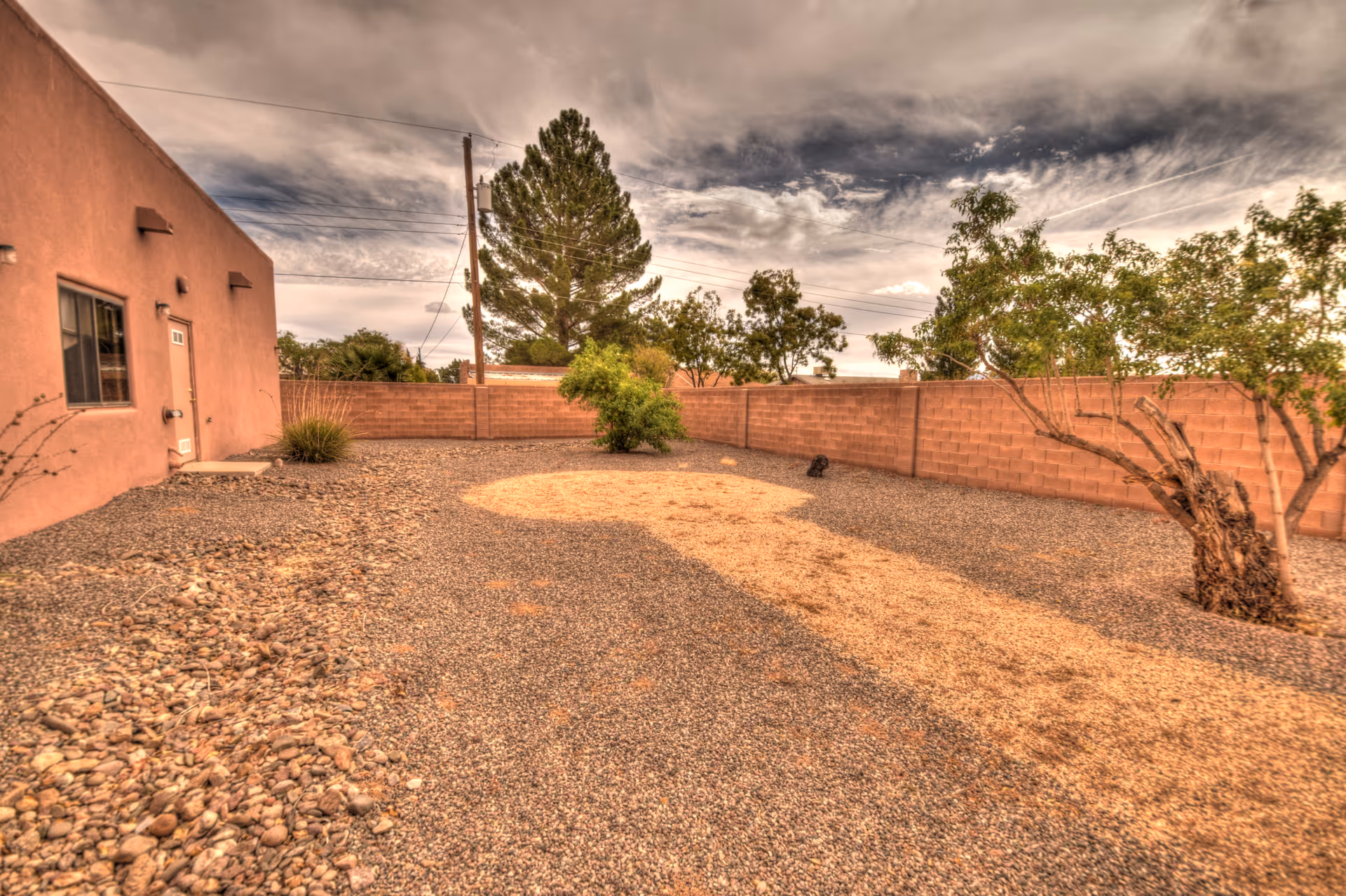 A fenced outdoor area with gravel ground and a few small trees and bushes. The area is bordered by a brick wall and the side of a building with a window and a door. The sky is cloudy with a mix of light and dark clouds.