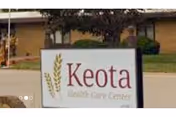 Outdoor view of a sign for Keota Health Care Center with a building and some greenery in the background.