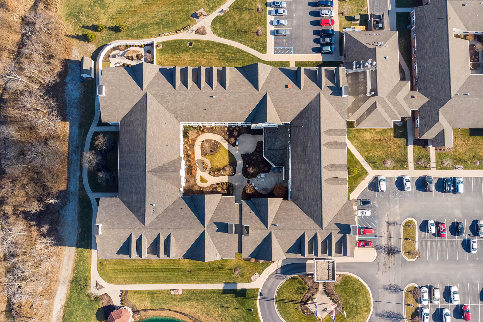 Aerial view of StoryPoint Fort Wayne West senior living facility showing the building with a central courtyard, surrounding green lawns, parking lots with cars, and nearby trees and pathways.