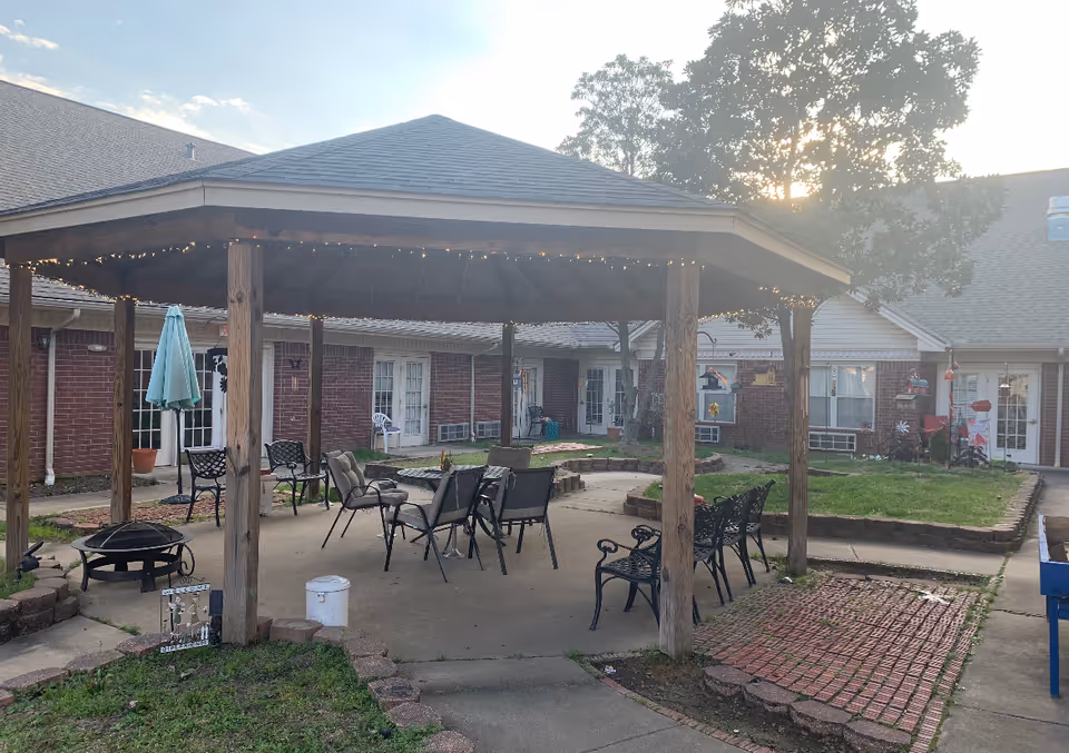 Outdoor patio area at Cypress Place featuring a wooden gazebo with string lights, several chairs and benches arranged underneath, a fire pit, and surrounding brick and concrete pathways with grass and trees in the background.