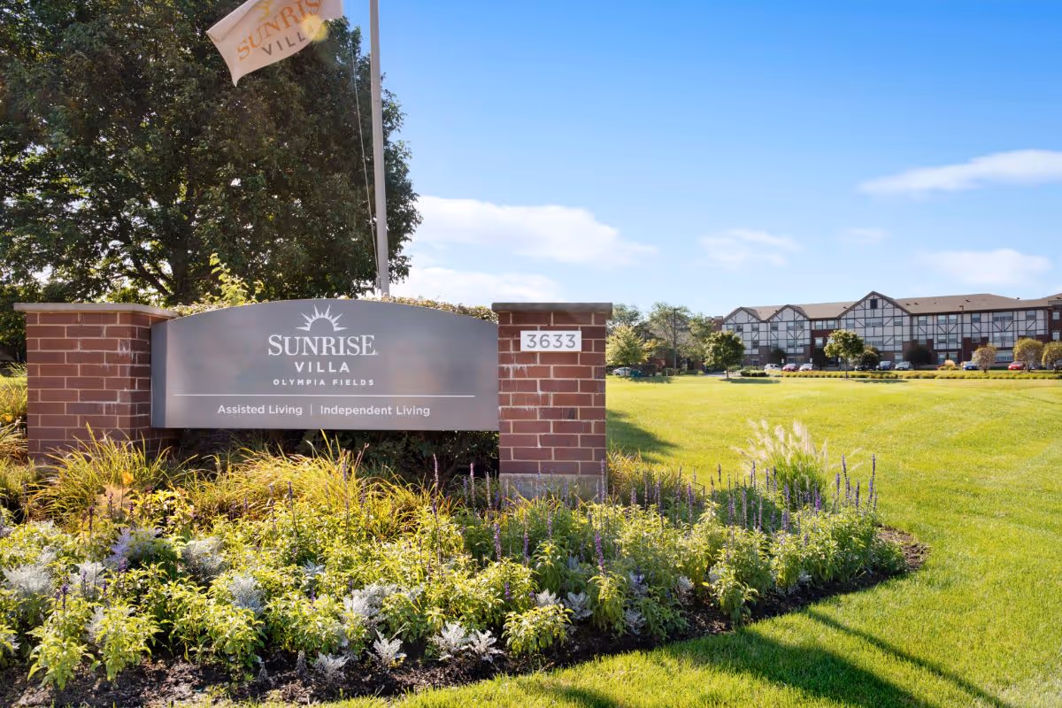 Outdoor view of Sunrise Villa Olympia Fields assisted living and independent living facility sign with a brick base and landscaped flowers in front. The building and parking lot are visible in the background under a clear blue sky.