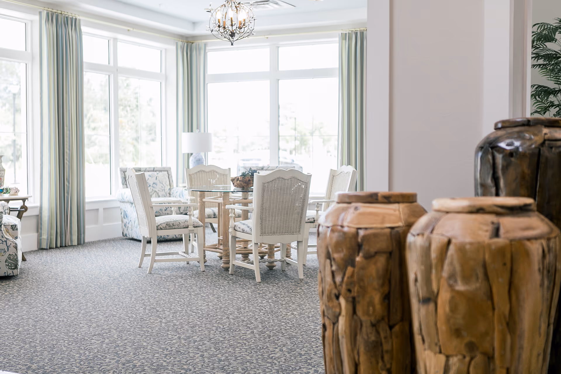 Bright and airy room with large windows covered by light green curtains, featuring a round glass-top table surrounded by white wooden chairs with patterned cushions. The room has a patterned carpet and decorative wooden vases in the foreground.