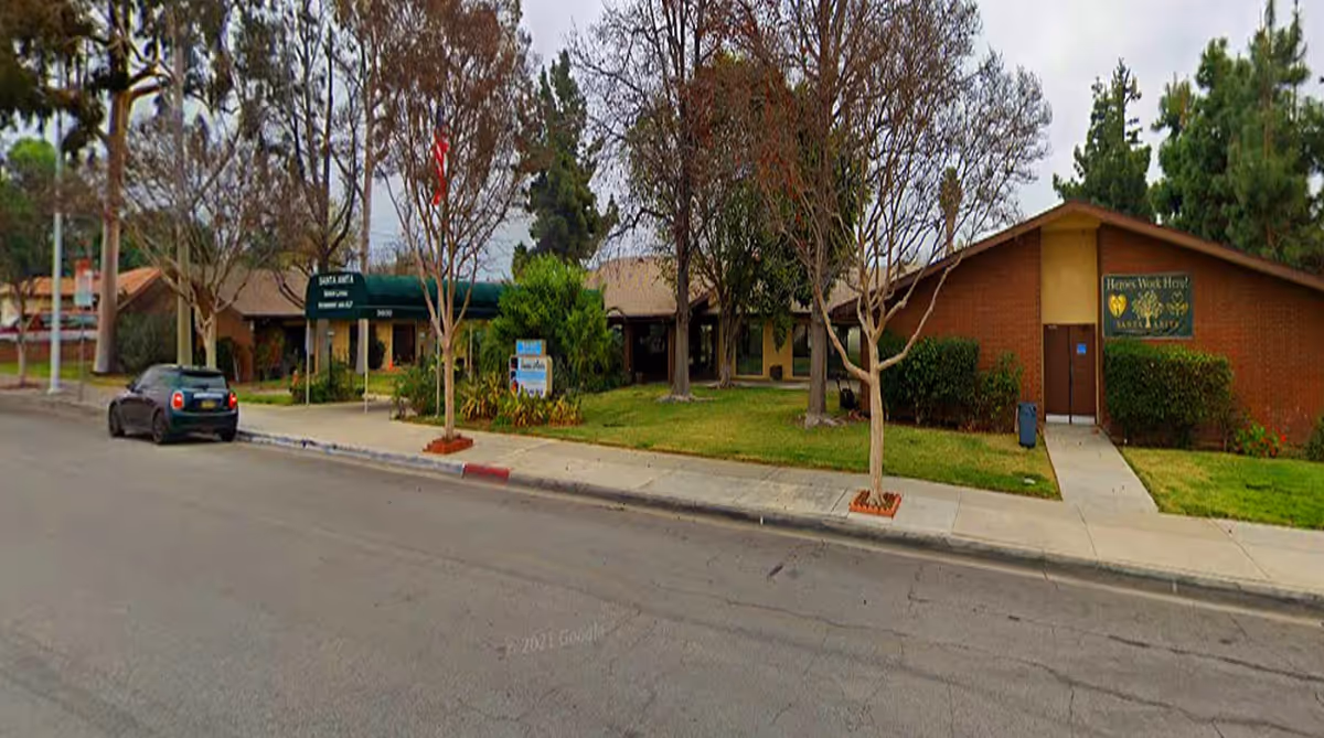 Exterior view of Santa Anita Assisted Living facility showing a single-story brick building with a green awning over the entrance, surrounded by trees and grass. There is a sidewalk and a parked car along the street in front of the building.