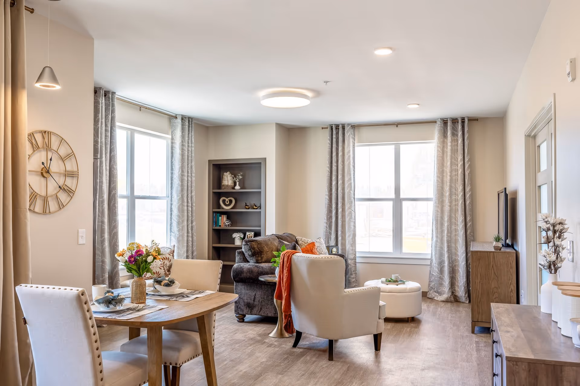 A bright and cozy living room and dining area with large windows covered by patterned gray curtains. The living room features a dark gray sofa, a beige armchair with an orange throw, a white round ottoman, and a wooden bookshelf with decorative items. The dining area has a wooden table set with two beige upholstered chairs, a vase with colorful flowers, and place settings. The room has light-colored walls and wood flooring, with modern ceiling lights and a large decorative wall clock.