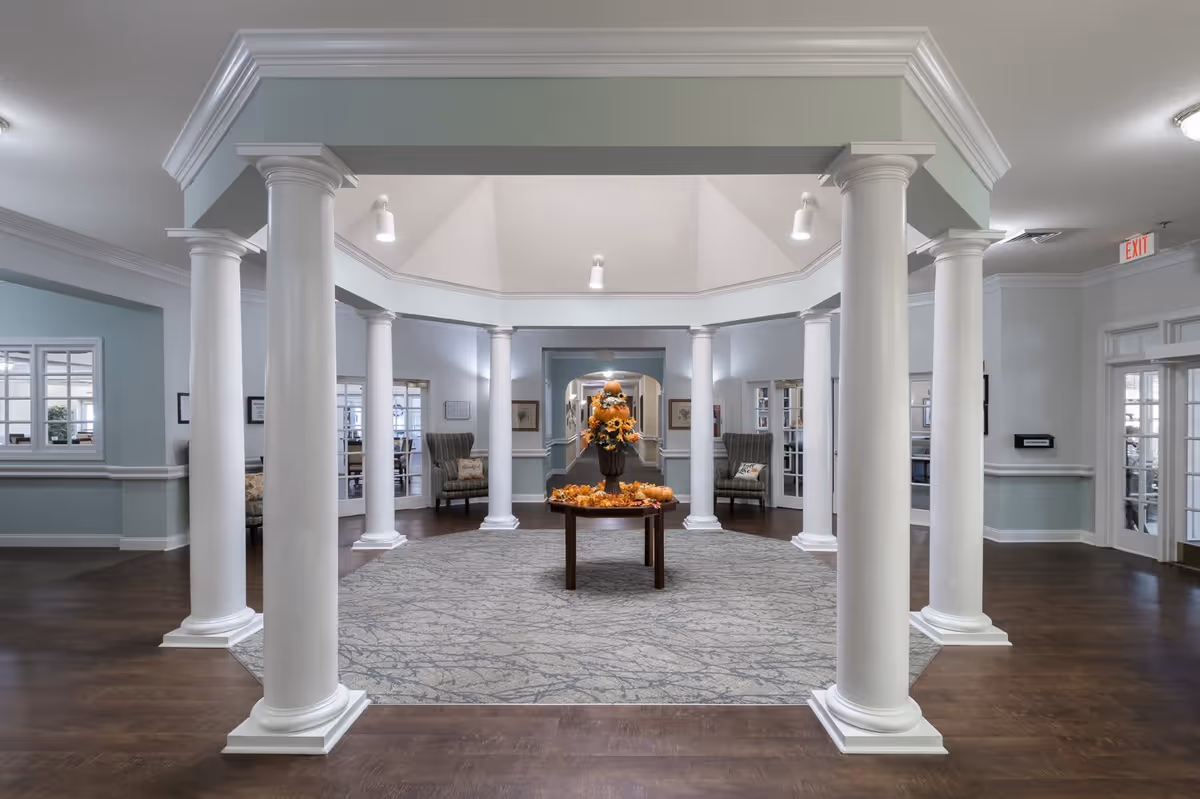 Interior view of a senior living facility lobby area with white columns surrounding a central carpeted space. In the middle, there is a round wooden table decorated with a fall-themed floral arrangement. The walls are painted light blue with white trim, and there are seating areas with armchairs and framed pictures along the walls. The ceiling is vaulted with recessed lighting.