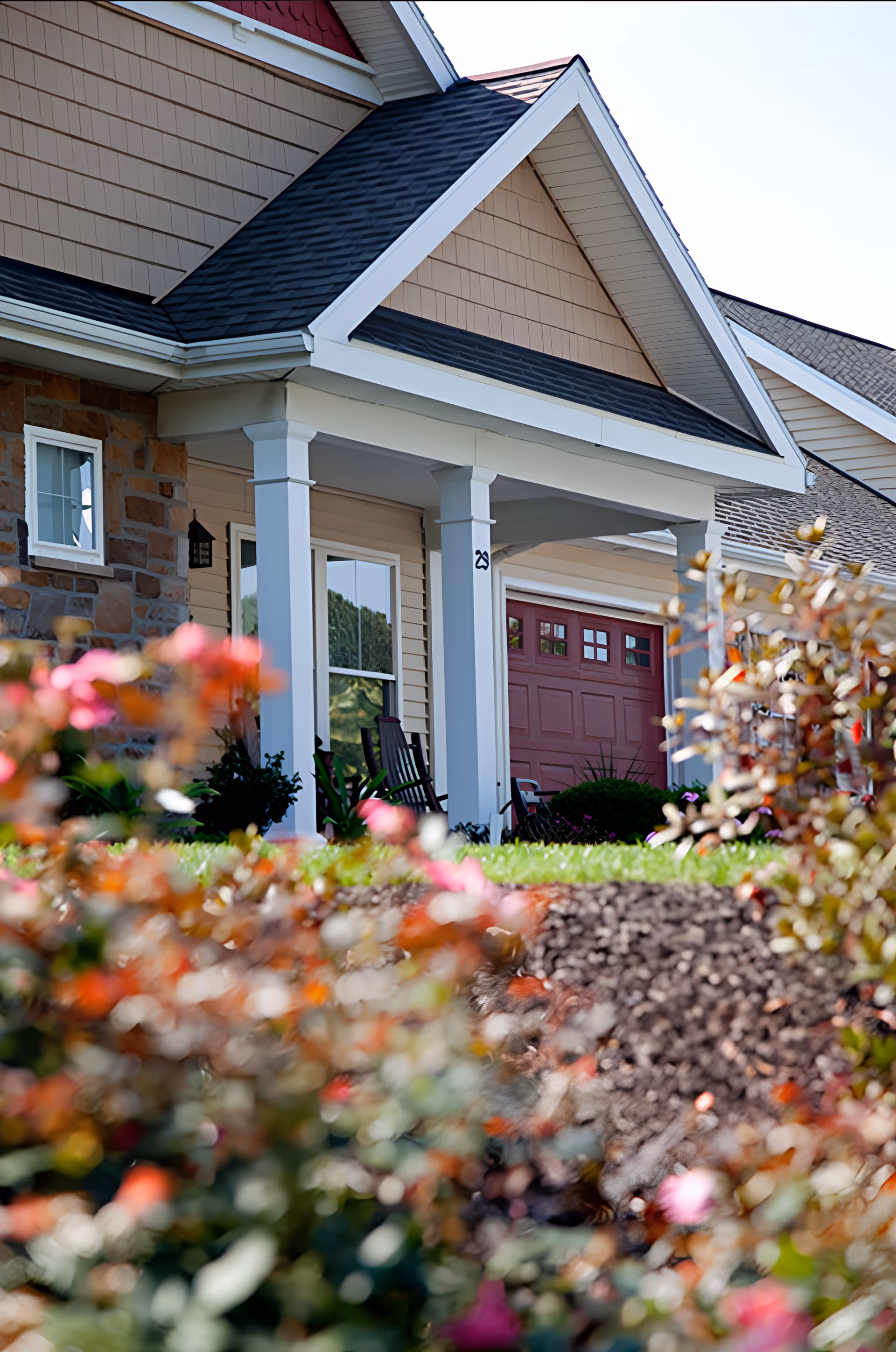 Front exterior view of a residential-style building with a covered porch supported by white columns, a red garage door, and a garden with colorful flowers in the foreground.
