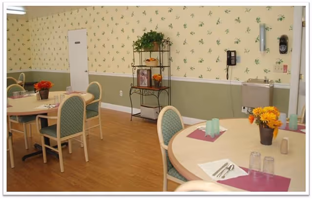 Small dining room with round tables set with placemats and flowers, upholstered chairs, a wall-mounted phone and water fountain, and a decorative shelving unit against wallpapered walls.