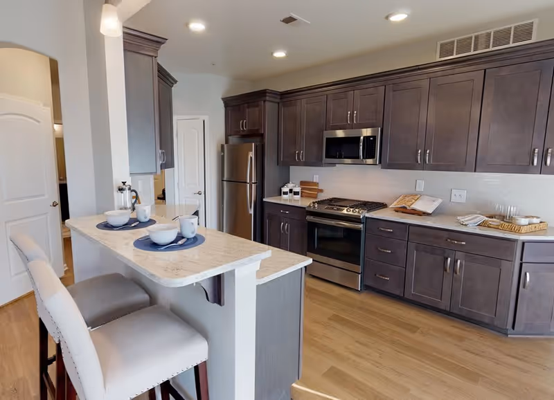 Modern kitchen with dark wood cabinets, stainless steel refrigerator, stove, and microwave. A white countertop island with two beige cushioned bar stools is set with bowls and cups on blue placemats. The kitchen has light wood flooring and white walls.