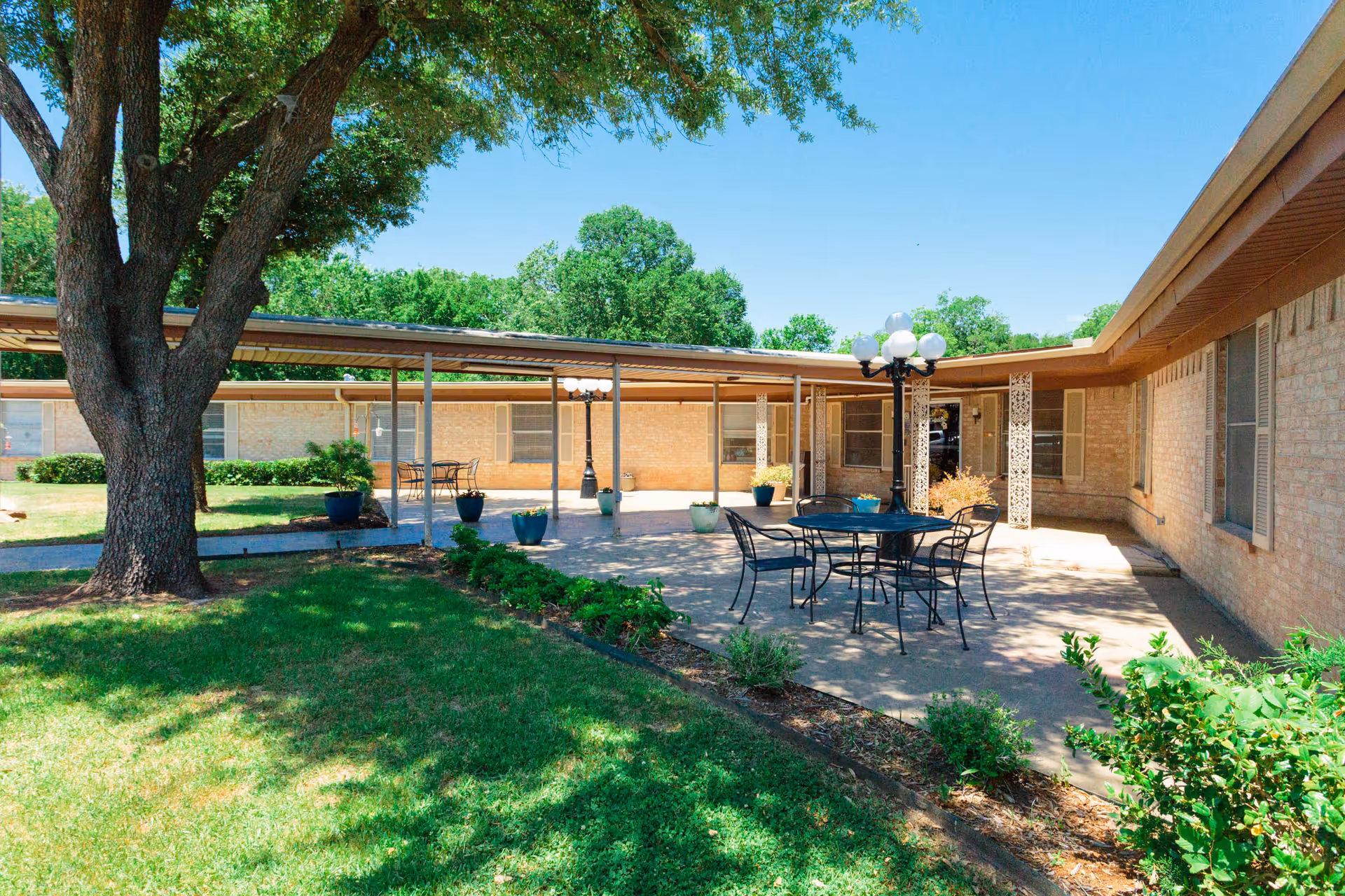 Outdoor courtyard area with a large tree, green grass, and a paved patio featuring a round metal table with four chairs and a decorative lamp post. The courtyard is surrounded by a single-story brick building with windows and a covered walkway.
