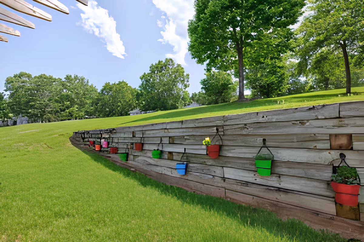 Grassy outdoor hillside with a wooden retaining wall decorated with colorful hanging planters and trees under a blue sky.