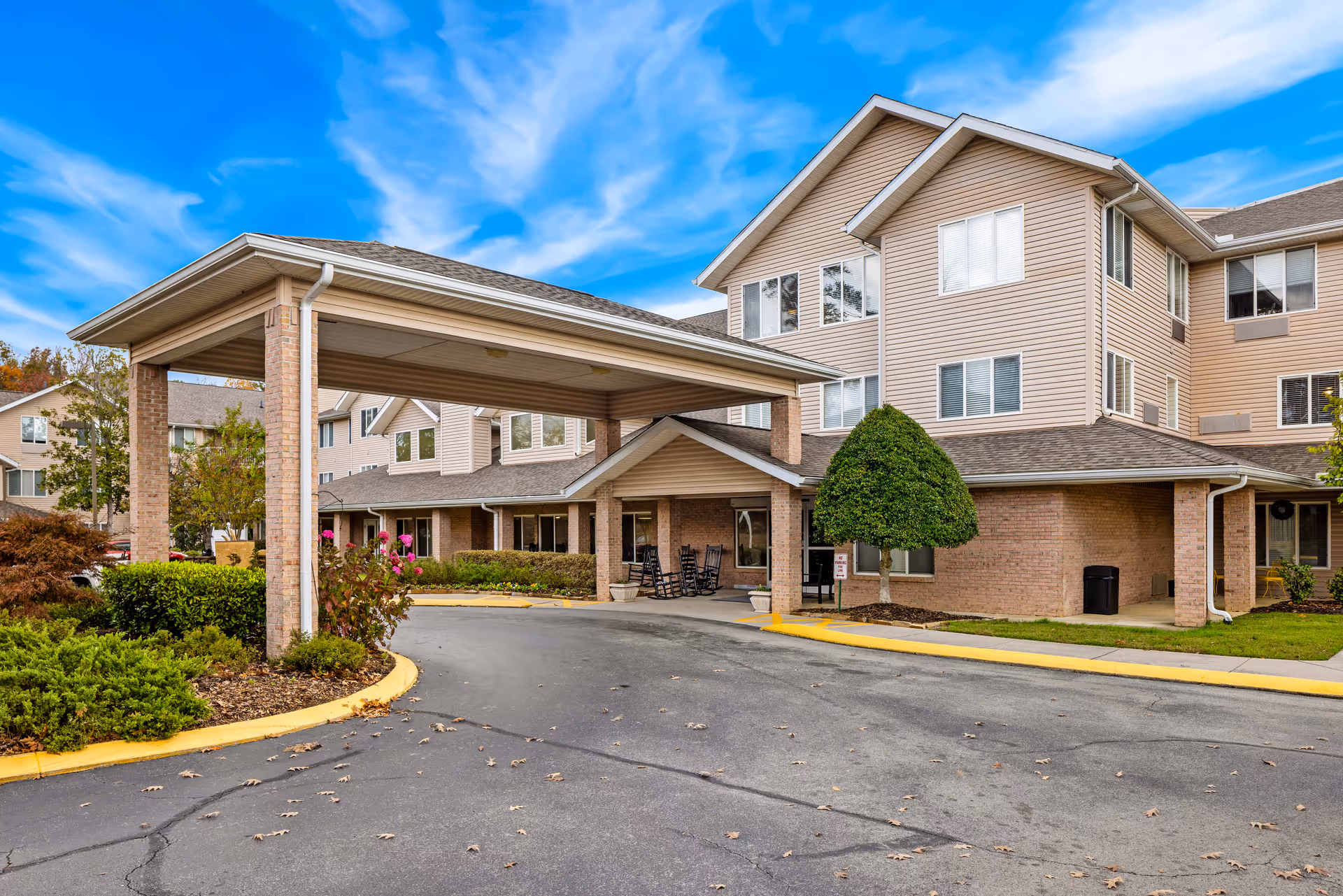 Exterior view of Creekside at Shallowford, a senior living community building with a covered entrance driveway, surrounded by landscaping including bushes and a shaped tree, under a blue sky with some clouds.
