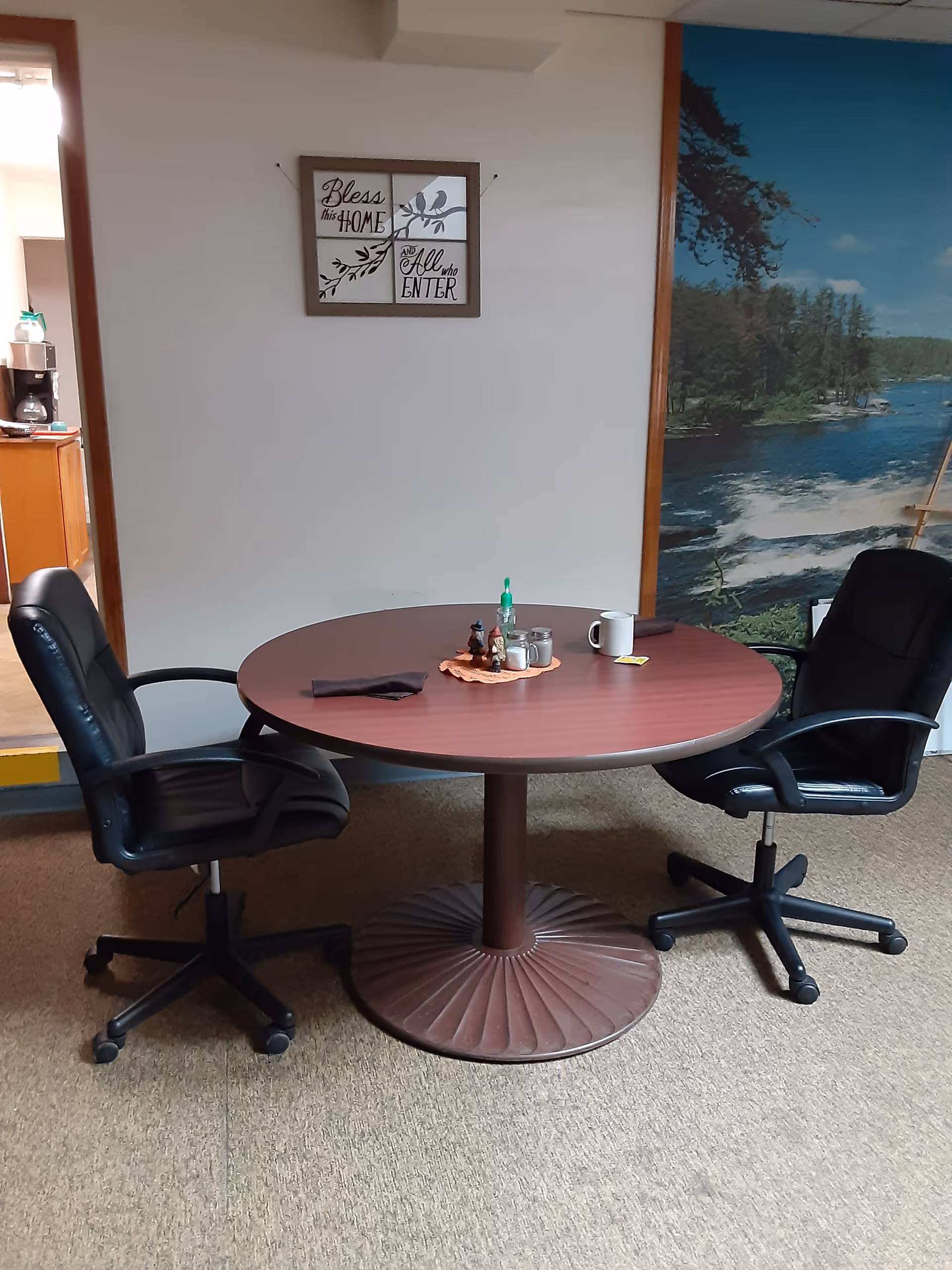 A round wooden table with two black office chairs on either side. On the table are a few items including a bottle of hand sanitizer, salt and pepper shakers, a coffee mug, and a napkin. Behind the table is a wall with a framed sign that reads 'Bless this HOME and All who ENTER'. To the right, there is a large wall mural depicting a river and trees.