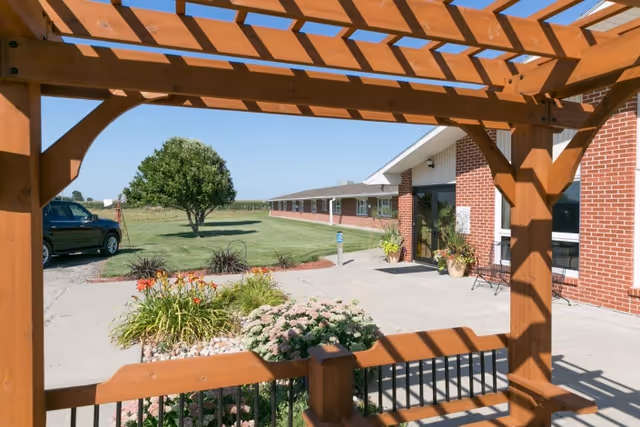 View from under a wooden pergola looking out towards the entrance of a single-story brick building with a concrete walkway, landscaped flower beds, a tree, and a parked black vehicle on a gravel driveway under a clear blue sky.