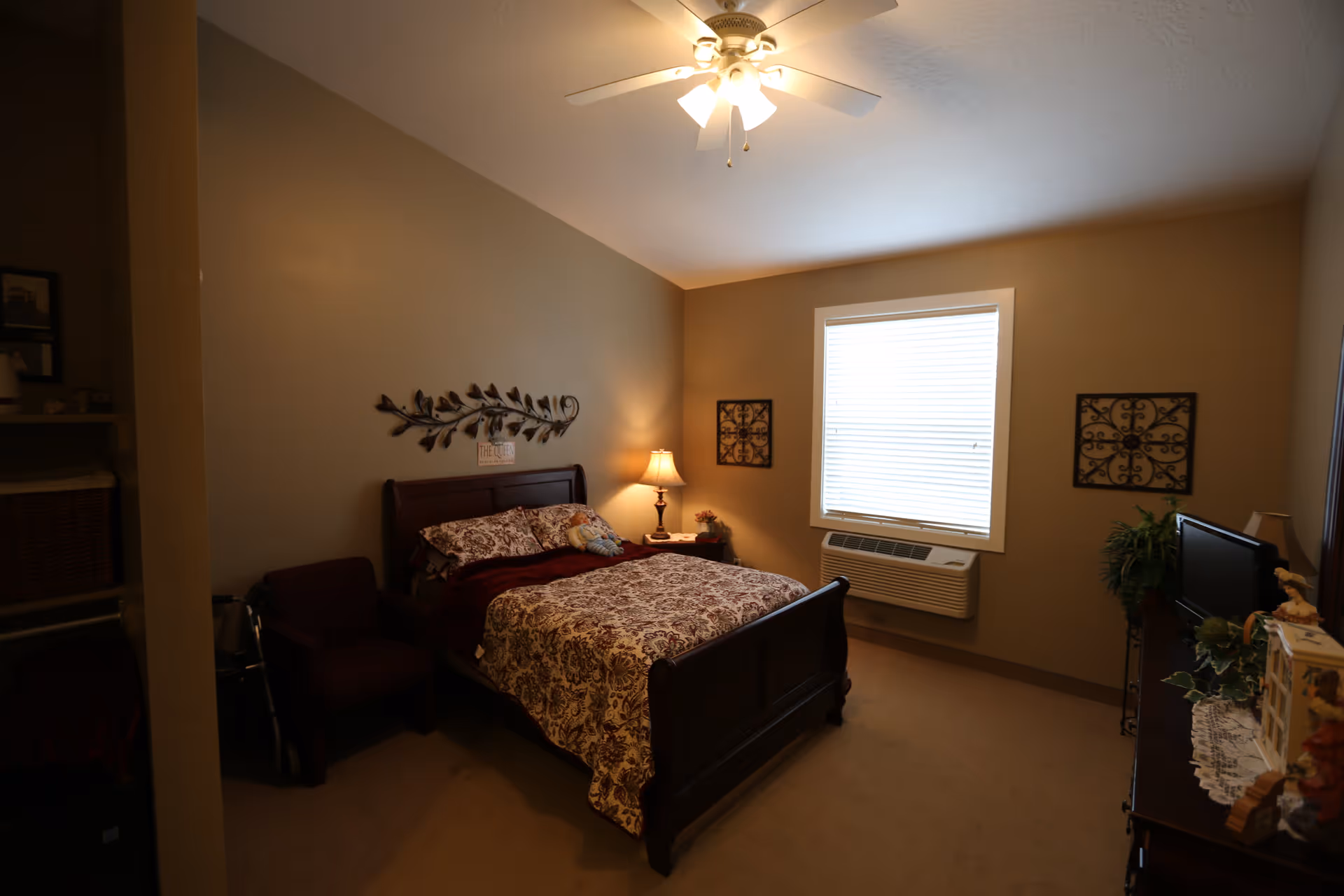 A cozy bedroom with a wooden bed frame, floral patterned bedding, a ceiling fan with lights, a window with blinds, a nightstand with a lamp, a small armchair, a walker, decorative wall art, and a dresser with a TV and plants.