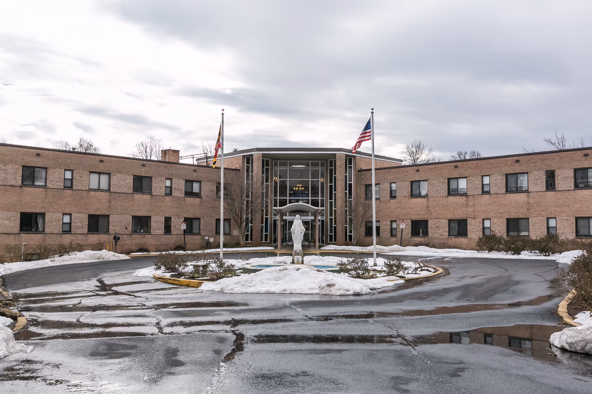 Front exterior view of a two-story brick nursing and rehabilitation facility with a circular driveway, two flagpoles with flags, a statue in the center, and patches of snow on the ground under a cloudy sky.