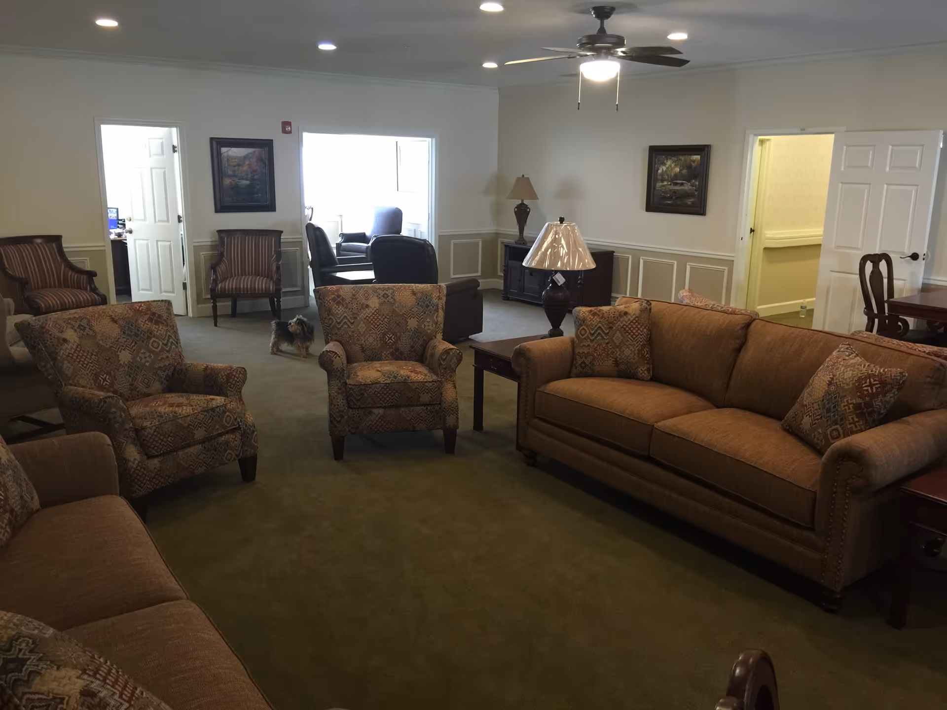 Common room in a senior living facility with sofas and patterned armchairs arranged around side tables, a ceiling fan, and a small dog on the carpet.